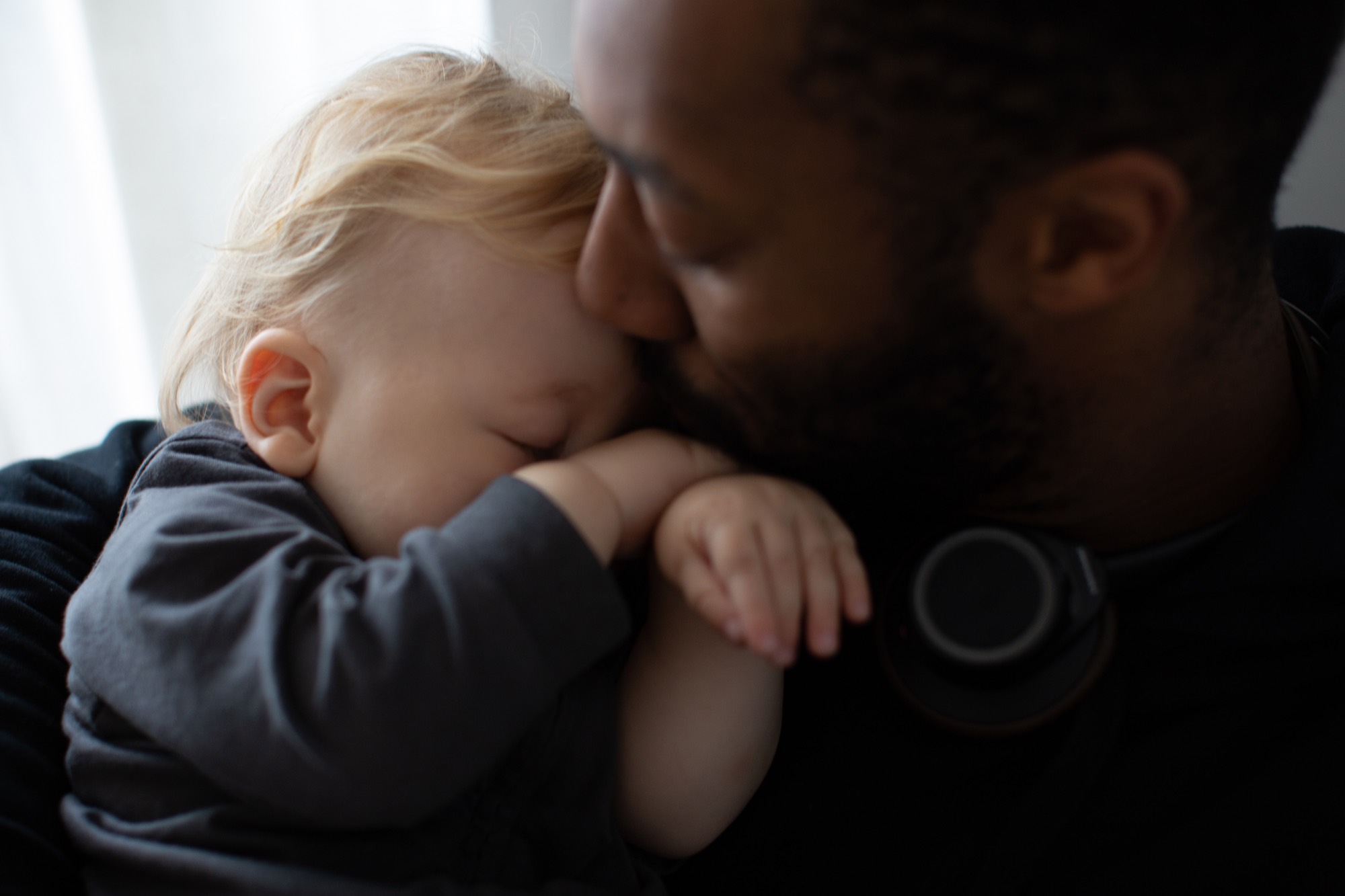 A father kisses his sleeping baby by the window.
