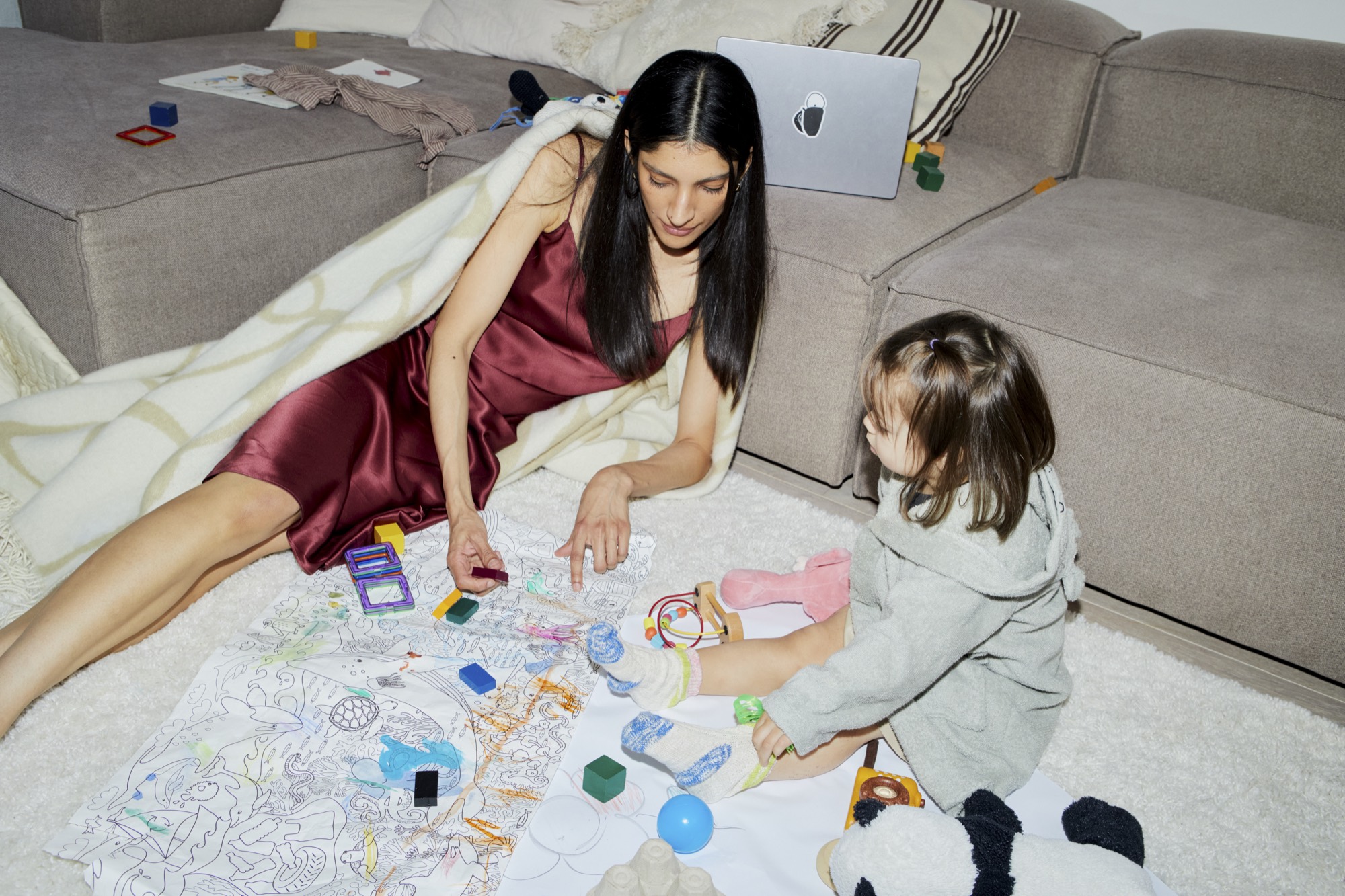A mother in a burgundy dress plays with her child on the living room floor.